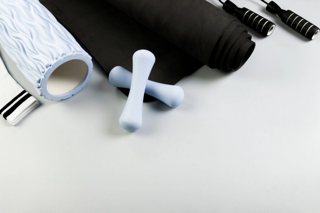 Top view of blue dumbbells, blue massage roller, black jumping rope and rolled yoga mat on grey background. White sport socks, fitness, sport, gym and health.