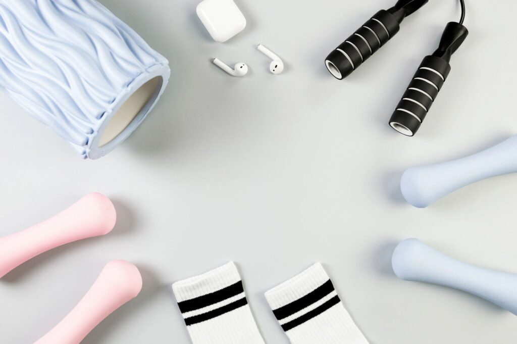 Top view of pink dumbbells, blue dumbbells, blue massage roller, black jumping rope and white earphone case on grey background. White sport socks, fitness, sport, gym and health.